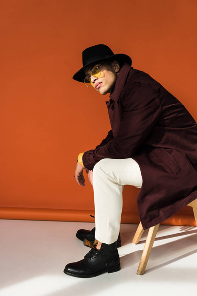 stylish mixed race man in hat and sunglasses sitting, looking at camera and posing on orange - 写真・画像