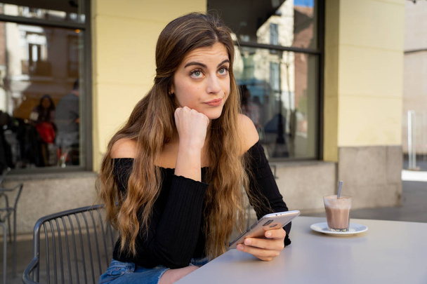 Pretty young woman with stylish black top, looking bored drinking coffee, sending messages on smart phone outside at coffee shop. Feeling tired and dull. In lifestyle and technology use concept. - Foto, Bild