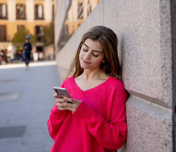 Pretty happy student woman chatting online on smart phone using social media apps outside city street. Beautiful teenager wearing stylish clothes feeling free. People, lifestyle and mobile use. - Photo, Image