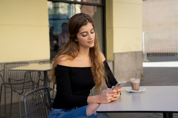 Pretty stylish young woman happy drinking a coffee, sending messages on mobile smart phone outside at a coffee shop. Feeling relaxed and carefree. In lifestyle and technology concept. - Fotografie, Obrázek