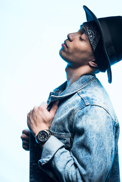 side view of handsome stylish mixed race man in denim and hat posing isolated on blue - Foto, immagini