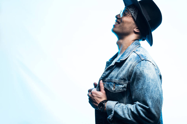 side view of handsome mixed race man in denim and hat posing on blue with copy space - Фото, зображення