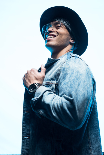 handsome smiling mixed race man in denim and hat posing on blue - Fotó, kép