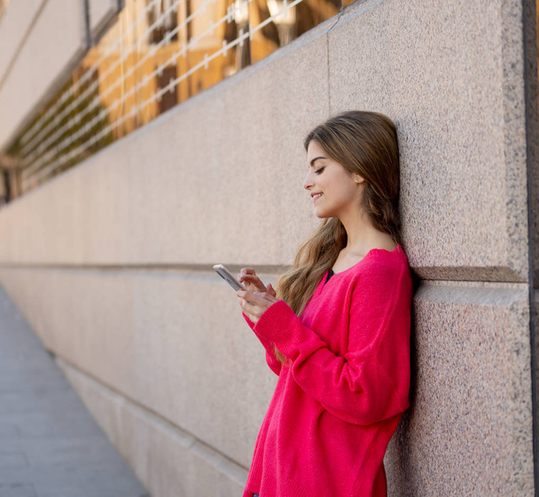 Redelijk tevreden student vrouw online chatten op slimme telefoon met behulp van sociale media apps buiten stad street. Mooie tiener dragen van stijlvolle kleding vrij gevoel. Mensen, levensstijl en mobiel gebruik. - Foto, afbeelding