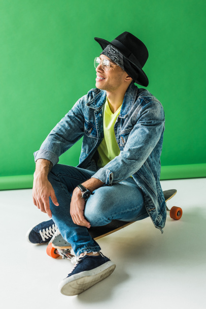stylish mixed race man in denim sitting on skateboard and smiling on green - Φωτογραφία, εικόνα