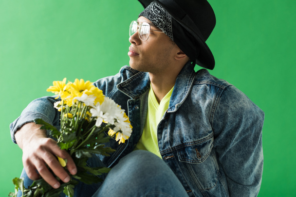 stijlvolle mixed race man in denim poseren met bloemen op groen scherm - Foto, afbeelding