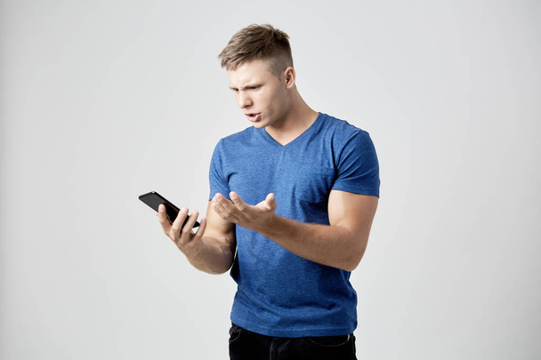 The guy dressed in blue t-shirt looks astonished at his mobile phone - Foto, imagen