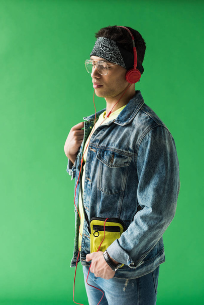handsome mixed race man in headphones and denim listening music on green screen - Foto, immagini