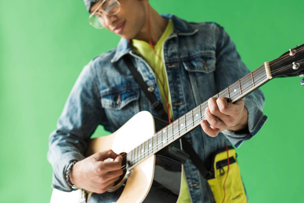 selective focus of mixed race man in denim playing acoustic guitar on green screen - Fotografie, Obrázek
