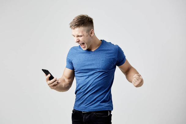 Ecstatic guy dressed in blue t-shirt looks at his mobile phone - 写真・画像