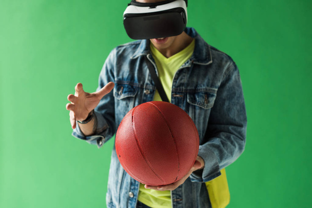hombre de raza mixta en los auriculares de realidad virtual gesto mientras sostiene el baloncesto en la pantalla verde
 - Foto, Imagen