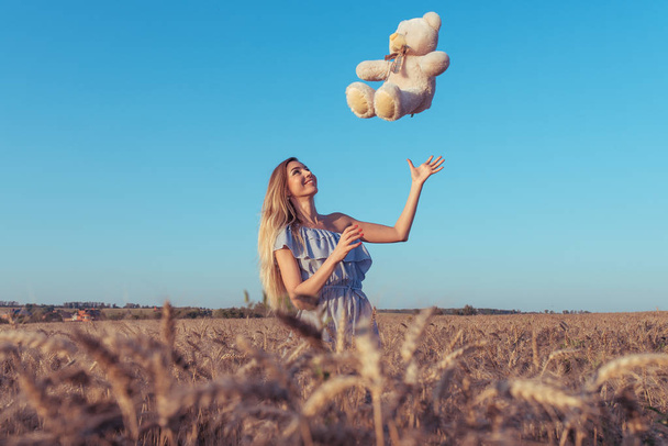 A girl in a dress, in a wheat field summer, throws up a toy teddy bear. Happy playing and having fun. Long hair with tanned skin. Free space. - Foto, Bild