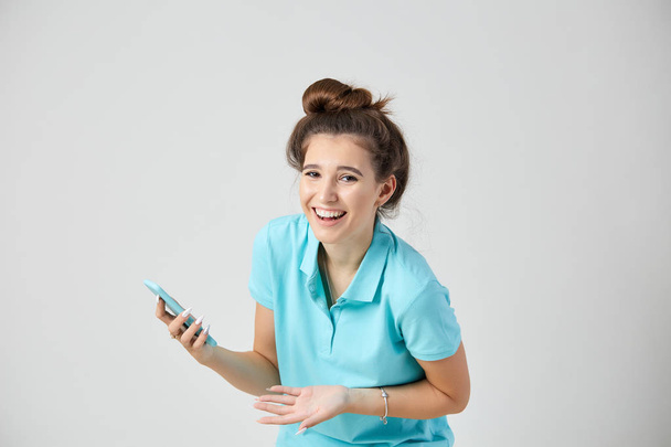 Joyful girl dressed in light blue t-shirt smiles and keeps mobile phone in her hand - Φωτογραφία, εικόνα