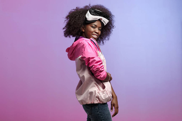 Funny curly brown-haired girl dressed in the pink sports jacket is wearing on her head the virtual reality glasses in the studio on neon background - Foto, Imagem