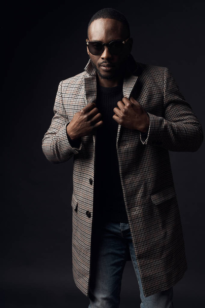 Serious cool man in dark sunglasses, black sweater and brown plaid coat and jeans. Studio, dark key - Fotó, kép
