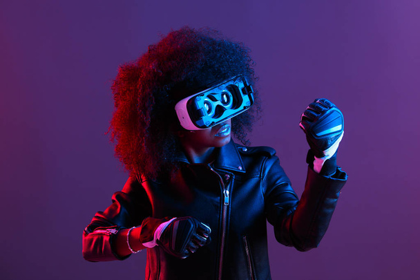Curly dark haired girl dressed in black leather jacket and gloves uses the virtual reality glasses on her head in the dark studio with neon light - Foto, Bild