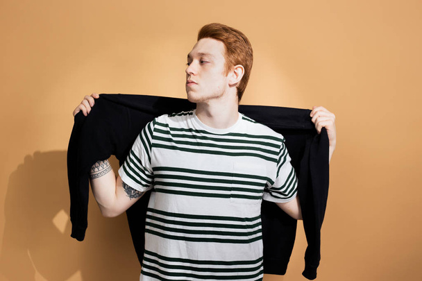 Stylish red-haired guy in a striped shirt with tattoo on his hand is posing with black sweater on his shoulders on the beige background in the studio - Φωτογραφία, εικόνα