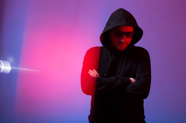 Portrait of fashion man in a black sweater with a hood and sunglasses in neon light in the studio - Fotografie, Obrázek