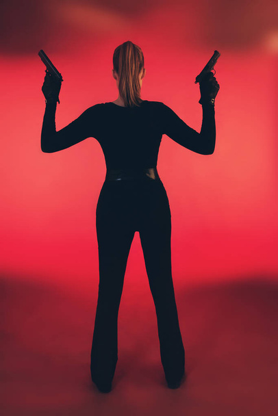 Girl with two guns dressed in black. Standing against red backgr - Fotografie, Obrázek