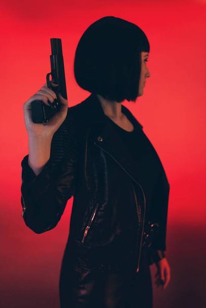 Woman with gun dressed in black against red background. - Фото, изображение
