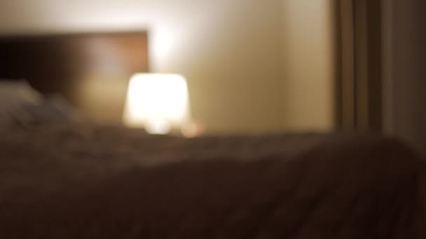 Imagen interior de la sala de noche con cama doble grande cubierta con colcha e iluminada por la luz de la lámpara amarilla que cambia lentamente de borrosa a enfocada
 - Metraje, vídeo