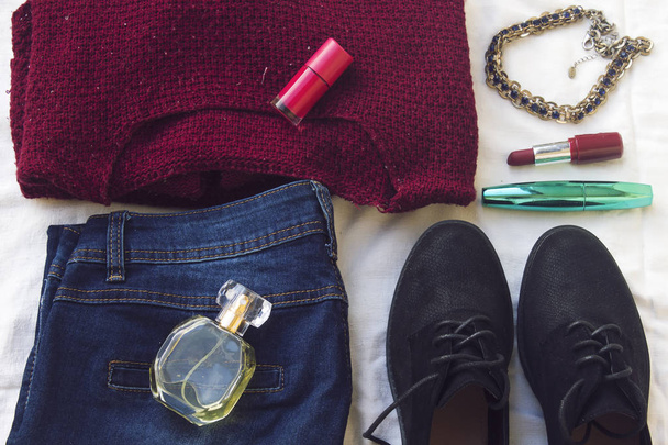 blue jeans, red oversize knitted sweater and black flat shoes, necklace and pink nail polish, mascara, bottle perfume on white sheet. Overhead view of woman's casual day outfits. Trendy hipster look. - Photo, Image