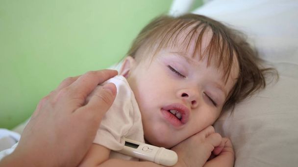 мама вимірює температуру з термометром малюкові і пестить дитину рукою. крупним планом. дитина спить в лікарняному палаті на білому ліжку
. - Фото, зображення