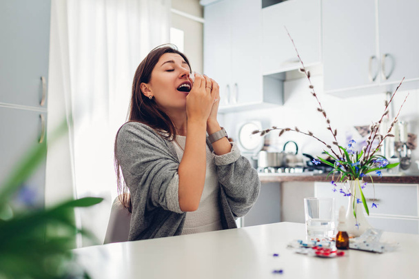Весенняя аллергия. Молодая женщина чихает из-за цветов, окруженных таблетками на кухне. Сезонная аллергия
. - Фото, изображение