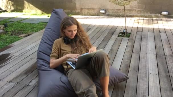 young woman working on laptop while sitting on beanbag chair at wooden terrace  - Záběry, video