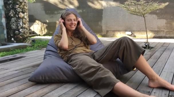 atraktivní žena v příležitostném stylu poslouchat hudbu ve sluchátkami a relaxaci, když sedí na židli s fazolovou brašnou na dřevěné terase  - Záběry, video