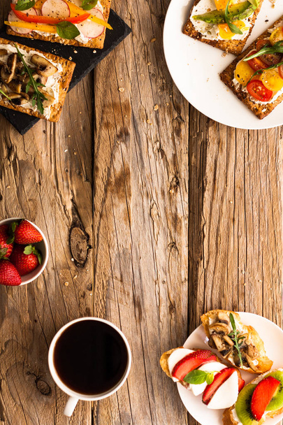 Ontbijttafel met smakelijke bruschetta crostini toast, roomkaas, verse groenten en fruit en een kopje zwarte koffie. Ochtend Heide uitgebalanceerd dieet. Kleurrijke ingrediënten op rustieke houten achtergrond - Foto, afbeelding