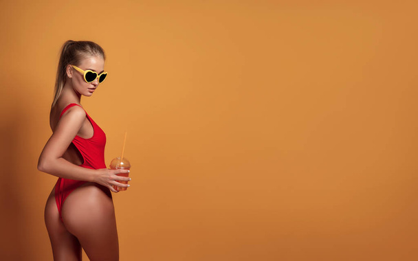 Lovely young female in swimwear and sunglasses holding glass of fresh drink while standing on yellow, orange hot background - Foto, Bild