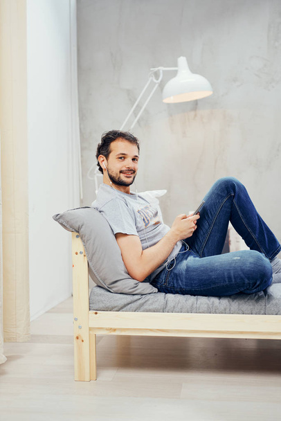 Uomo arabo sorridente seduto sul letto in camera da letto e denunciare smart phone. In cuffie di orecchi
. - Foto, immagini