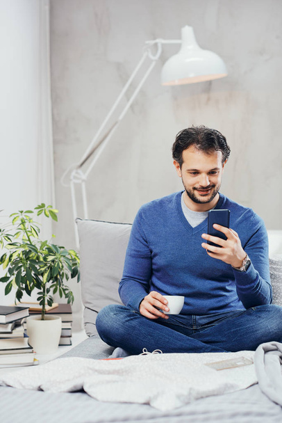 attrayant sourire arabe habillé occasionnel assis sur le lit dans la chambre, boire du café et en utilisant un téléphone intelligent pour envoyer des SMS
. - Photo, image