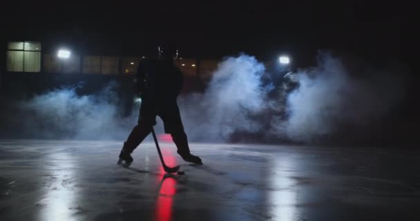 Male hockey player with a puck on the ice arena shows dribbling moving directly into the camera and looking directly into the camera against a dark background in the smoke - Video