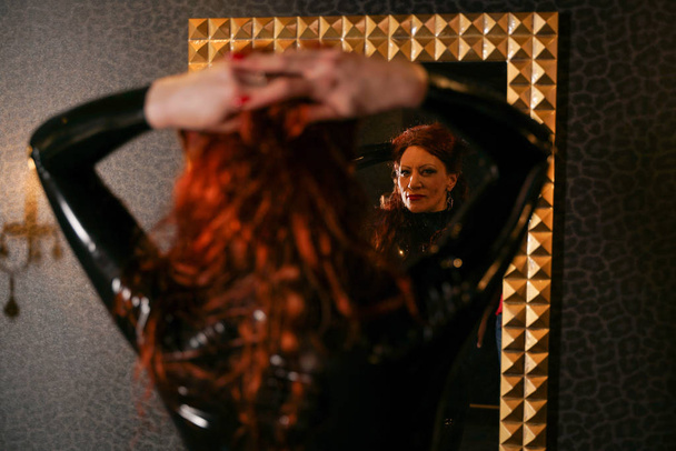 seksuele fetisj roodharige vrouw dragen van zwarte latex rubber catsuit en kijken naar de spiegel in de donkere kamer - Foto, afbeelding