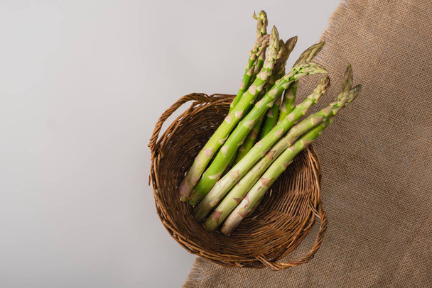 top view of green asparagus in wicker basket near sackcloth on grey background - Фото, изображение