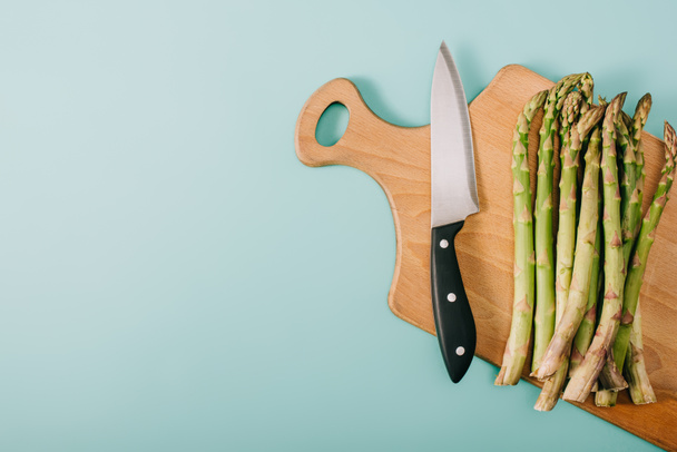 top view of green raw asparagus on wooden cutting board with knife on blue background - Foto, Imagen