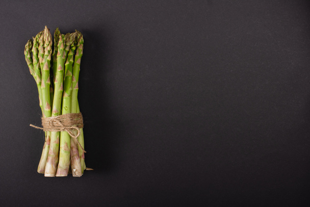 top view of bunch of green uncooked asparagus tied with rope on black background - 写真・画像