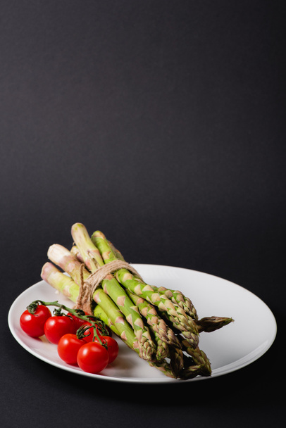 bunch of green uncooked asparagus tied with rope and cherry tomatoes on white plate on black background - Фото, зображення