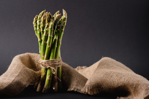 bunch of green raw asparagus tied with rope near sackcloth on black background - Photo, Image
