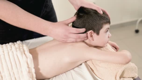 A male physiotherapist masseur makes a healing relaxing massage to a little smiling boy lying on a massage bed. Head massage - Video