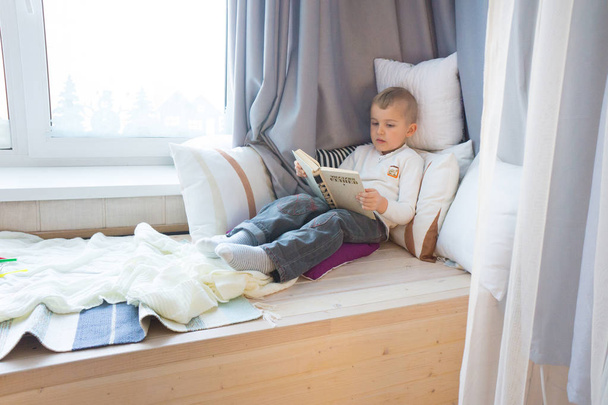 The boy is reading a book near the window. Winter. Pillows aroun - Valokuva, kuva