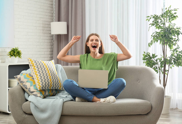 Emotional young woman with laptop celebrating victory on sofa at home - Foto, Imagen