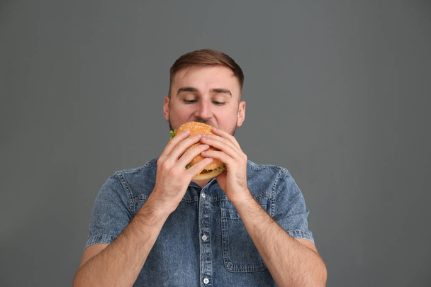 Young man eating tasty burger on grey background - 写真・画像