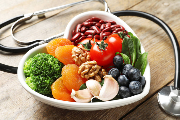Bowl with products for heart-healthy diet and stethoscope on wooden table - 写真・画像