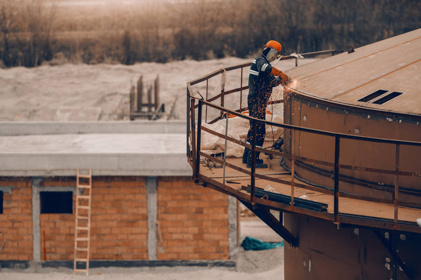 Werknemer in beschermende uniform en helm slijpen top van de toren op de bouwplaats. - Foto, afbeelding