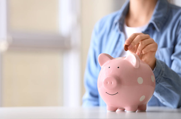 Woman putting coin into piggy bank at table indoors, closeup. Space for text - Photo, Image