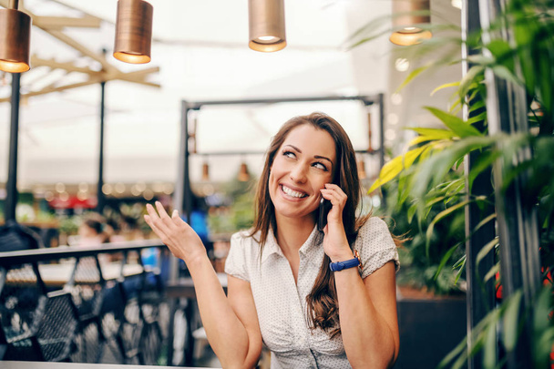 Jonge Kaukasische glimlachend brunette zitten in Cafe en met behulp van slimme telefoon voor gesprek. - Foto, afbeelding