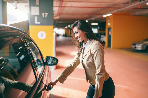 Smiling attractive brunette dressed smart casual opening car door at public garage. - Foto, imagen